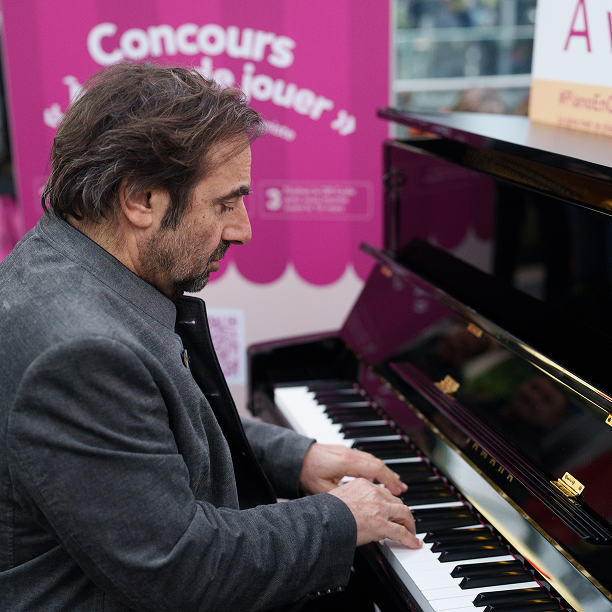 La photo montre André Manoukian, le parrain du dispositif Piano En Gare, en train de jouer sur le piano installé à Paris Gare de Lyon
