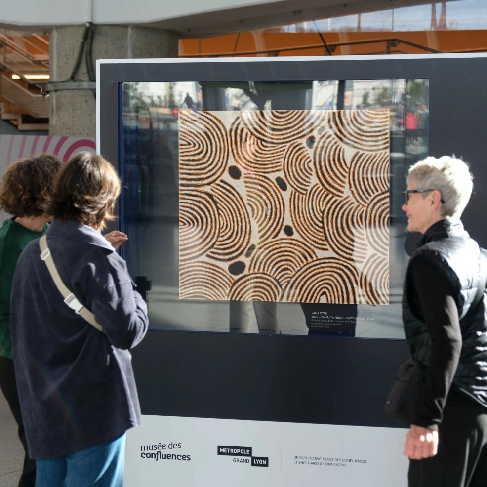 Deux femmes ainsi qu'une médiatrice se tiennent devant une œuvre originale exposée en gare de Lyon Part Dieu dans le cadre de l'opération "Un jour, Une œuvre". Une toile aborigène issue du musée des Confluences se trouve dans une cimaise.