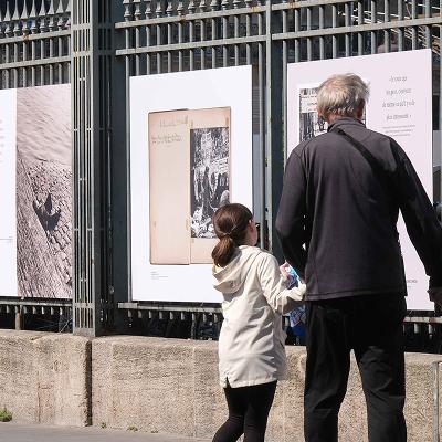 Un grand-père et sa petite fille découvrent une exposition installée sur les grilles extérieures de Paris Et. On y voit des photographies, des citations, des documents d'archive.