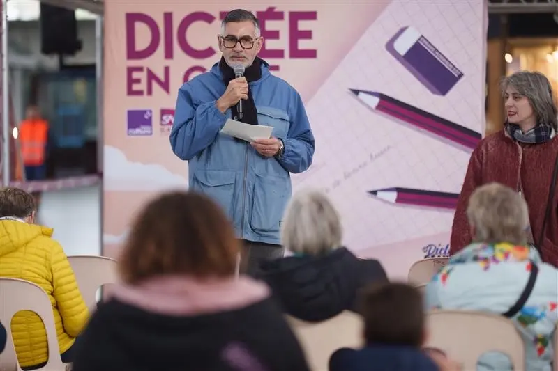 La scène se déroule en gare de Lille Europe, où se tient une dictée géante. On voit des personnes de tous âges en train d'écrire le texte dicté par l'animateur de l'événement, Rachid Santaki